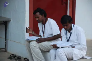 Two young men in white ashram clothing sit on a concrete step in front of a red-framed doorway against a pale wall. The man on the left wears a white t-shirt, light trousers, a dark rudraksha-style mala and a pendant, and holds open printed pages while gesturing with his right hand. The man on the right wears a white kurta and white trousers with a blue beaded mala, and reads from an open booklet. Black sandals rest on the ground at the lower left, and a small grey table edge is visible at the left.