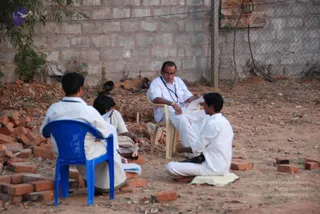 Outdoor photo of a small group sitting informally on a dusty ground beside a brick wall and chain-link fence. A bald man with glasses, wearing a white shirt and white dhoti with a lanyard, sits on a beige plastic chair holding papers and speaking. Facing him, a young man in white kurta and dhoti sits cross-legged on a white cloth on the ground. On the left, a man in white sits on a blue plastic chair, and a partially visible person in white sits nearby. Scattered red bricks, a small plant and debris surround them.