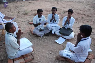 Outdoor photo of five men in white ritual clothes sitting in a loose circle on bare red earth, with low brick seats and dry grass around them. Most have palms pressed together in anjali mudra; the man at left holds an open book, the man second from left holds loose papers, and the man at right holds a small folded booklet. Several wear blue lanyards. A dark shoulder bag sits on the ground near the centre. A man at the far left edge, partly cropped, holds papers while sitting on a chair. No a person candidate is visibly present.