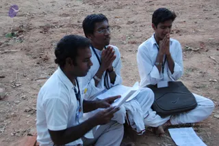 Three young men sit cross-legged on bare red earth outdoors, in plain white shirts and white trousers with lanyard badges around their necks. The man on the left holds an open booklet and reads, while the two beside him press their palms together in anjali mudra with eyes closed. The man on the right rests a black zippered bag and loose papers beside him. Small dry leaves and sparse grass lie on the ground, with a purple logo in the upper left corner.