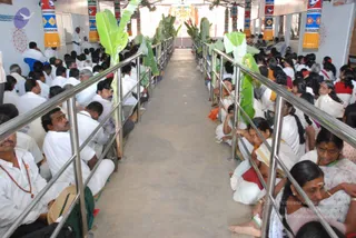 A long hall with a central aisle lined by stainless steel railings and banana plants with green leaves. Rows of seated devotees fill both sides, mostly men and women dressed in white, with some in saffron, red and cream saris. A man in white stands at the far left near the wall, and a man in a red-trimmed shirt sits with rudraksha beads visible on a figure at the left. Colourful striped pillars with floral patterns line the walls, and a decorated shrine area with a deity murti and garlands is visible at the far end. The scene suggests a seated congregation gathered for a ritual or satsang.