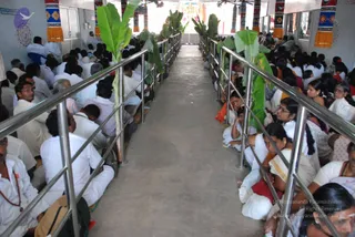A long central aisle runs between two rows of seated devotees in an indoor hall lined with banana plants tied to metal railings. Most people wear white clothes, and some men wear rudraksha malas. Women on the right wear white saris with coloured borders. At the far end a decorated altar area is visible above the crowd. Faint copyright and source text is overlaid across the lower centre of the photo.