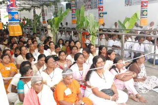A large seated crowd of devotees fills an indoor hall, with many women in white sarees and some in orange, pink and yellow garments sitting close together on the floor behind a metal railing. Several women wear rudraksha-style beads and white vibhuti marks on their foreheads, and a few wear name badges. Banana plants and decorated pillars in red, orange and white line the hall, and men in white shirts stand or sit toward the back. Faint printed signage is visible on the upper wall. A white rope barrier runs across the foreground.