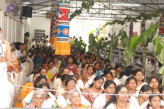 A large crowd of mostly women sits seated on the floor of a covered hall, many in white saris with vibhuti on their foreheads. Some wear orange, red and yellow saris, and several have red bindis and gold necklaces. A row of white-clad devotees is seated in front, with a white rope line separating the seated group from an aisle. Behind them, tall painted pillars in blue, red, green and yellow with floral and bird motifs frame the space, and banana plants with green leaves are placed along the edges. A few people stand at the left edge, one in an orange garment. Several people are visible at the back of the hall near a white doorway, and a loudspeaker hangs on the left wall.
