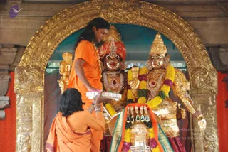 Photo inside a temple shrine framed by an ornate gold arch. A man with long dark hair and an orange saffron garment stands at left, a rudraksha-style garland at his neck, reaching toward a golden altar. Before him, a person in orange robes with dark hair holds a silver tray with lamps or offerings. Two large golden deity murtis stand behind the altar, one wearing a red and gold crown and jewelled necklaces, the other with a tall gold crown, both adorned with yellow and purple garlands. A golden kalasha-style vessel with a garland sits on the altar in front. Teal fabric hangs behind the shrine.