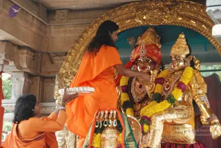 Outdoor temple shrine photo. A person in an orange saffron robe with long, swept-back dark hair stands at the center, reaching toward two golden deity murtis adorned with yellow and purple marigold garlands, rudraksha-style beads and red-and-gold headdresses. The left murti wears a red and gold crown-like headdress. The figure in orange appears to be performing abhishekam or puja. A second person in orange, partially visible at lower left with bangles, holds a silver plate or tray. Ornate golden arch frames the shrine, with stone pillars behind. Offerings of fruit and flowers sit at the base in front of the murtis.