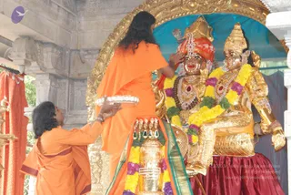 A photograph of a temple ritual at a shrine with ornate golden arches. Two figures in orange robes stand before a pair of golden deity murtis seated side by side, one wearing a red-and-gold crown and the other a gold crown with a red-gemmed necklace. The standing figure in long dark hair and an orange shawl reaches up to touch the head of the left murti with one hand. A second person in an orange garment kneels at the lower left, holding a silver plate with both hands toward the murtis. Yellow, purple and green garlands drape the deities and a brass kalasha in front of them, with a red-and-green cloth beneath. Red curtains hang at the left edge.