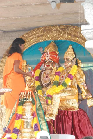 Photograph of a temple shrine with two gilded murti deities seated side by side under an ornate golden arch. The deity on the left wears a red and gold turban, rudraksha-style beaded necklaces and red-gem jewellery; the one on the right wears a tall golden crown and gold armour-like adornments. Both are heavily garlanded with yellow, purple and green flower garlands. In the foreground, a person in a long saffron-orange robe with long dark hair stands at left, holding pink flowers toward the figures. Below, a golden kalasha-style vessel draped with flower garlands and an orange-green silk cloth sits on a stand. A blue backdrop hangs behind the shrine.