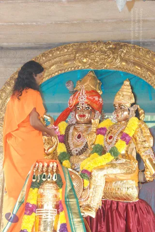 A photograph of a golden deity murti pair seated in a gilded arched shrine, the figure on the left wearing a red turban and jewelled necklaces with a red stone pendant, the figure on the right in a golden crown and garlanded in yellow and purple flowers with a maroon skirt. A long-haired person in an orange saffron robe stands at the left edge, reaching toward the murti and a brass or gold kalasha draped with a saffron and green-bordered cloth and yellow and purple garlands. A blue backdrop and ornate gold frame surround the shrine.