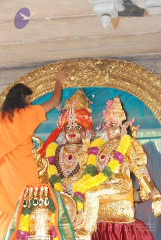 Photograph of a temple shrine with two gold-plated deity murtis seated under a large ornate gold arch against a teal backdrop. The murti on the left wears an orange-red turban and jewelled necklaces, and the one on the right wears a tall gold crown with pink flower petals and a dark red skirt; both are garlanded with yellow and purple flowers. A person in an orange robe with long dark hair stands at left, reaching up with one hand to the arch and scattering pink petals. In the lower left a brass kalasha-like vessel sits under an orange and green cloth with yellow and purple garlands, and falling petals are visible in the air.