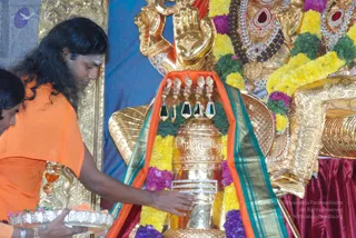 A man in an orange saffron robe with long, swept-back dark hair and a white-tilak forehead leans forward to pour liquid over a tall golden linga adorned with a gold and red-striped silk cloth. He holds a silver tray of offerings in his left hand. Behind the linga, a large golden deity murti wears a red and white jewelled necklace, with yellow and purple flower garlands draped around the figure. A second person with dark hair is partially visible at the left edge, and a red satin cloth covers the base of the altar. This appears to be a linga abhishekam ritual.