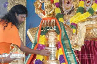 A photograph of a temple abhishekam at a golden deity. A man with long, swept-back black hair and a tilak on his forehead, wearing an orange saffron cloth draped over his shoulders, stands at left holding a silver tray with flowers and offerings in one hand while his other hand rests on a golden linga-shaped vessel. Behind the linga, a large golden deity murti wears a jewelled crown and necklaces, with yellow and purple flower garlands and a green-and-gold bordered orange cloth draped across it. A red satin cloth lies at right.