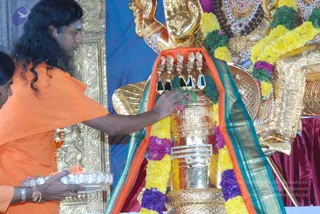 A photograph of a saffron-robed man with long, swept-back dark hair reaching toward a large golden linga on a golden deity figure. He wears an orange garment and has a silver tray held by a second person in orange at lower left, while a small figure with dark hair is partly visible at the left edge. The gold linga is adorned with yellow and purple flower garlands, green leaves, and a teal-and-orange cloth draped behind it. The scene suggests a temple puja setting with golden ornamentation and a maroon cloth at right.