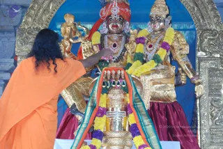 A photo of a temple ritual before a large gilded deity murti pair, shown in a decorated shrine with a blue backdrop and ornate gold arch. A figure in an orange saffron garment with long dark wavy hair stands at left, extending a hand to a silver-and-gold shivalinga-like object wrapped in an orange, green and teal cloth and draped with yellow and purple flower garlands. The seated deities are gold-coloured with red headgear, jewelled necklaces and yellow-and-green garlands, and the figure to the right wears a maroon skirt. Small gold lamps or offerings sit at the base.