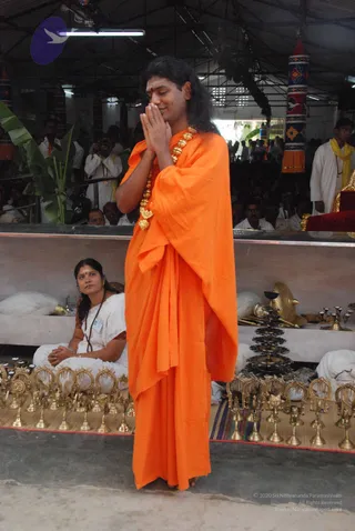 A standing man with long, swept-back dark hair and a red bindi on his forehead stands with palms pressed together in namaste, eyes lowered. He wears a flowing saffron-orange garment draped over his shoulders and a heavy ceremonial gold bead necklace. Behind him, a woman in a white sari with a rudraksha mala sits on the floor with hands folded in her lap, and rows of small brass bells and brass lamps are arranged on a mat in front. A tall tiered brass lamp stands nearby. A crowd of seated and standing devotees, many in white, fills the hall, with a red-cushioned seat and a yellow-shawled man visible at the right. A tall decorated pillar in red and white and a fan of green leaves are visible in the background.