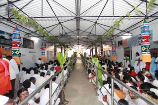 A photograph inside a long covered hall with a corrugated metal roof, decorated with green leaf garlands and tall painted cylindrical pillars in red, blue, yellow and orange. Many people sit cross-legged on the floor in white and light-coloured clothing, with a central aisle lined by steel railings and banana plants. Several standing figures in orange and saffron robes appear at the left, and women in saris stand at the right beside doorways. Framed posters and notice boards hang on the walls, and a small kolam-style red design is painted on the left wall.