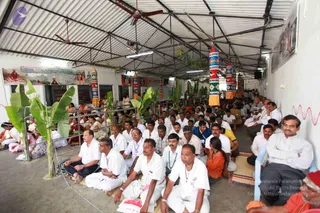 A large crowd of seated devotees fills a covered open-sided hall under a corrugated roof with ceiling fans. Most men wear white shirts and white dhotis, some with lanyards, seated on mats on the floor. Women in saris sit at left behind a metal railing beside potted banana plants. Tall cylindrical decorations with patterned red, white and blue panels line the hall. A framed poster on the left shows a red-robed figure and text, and a red wavy line is painted on the right wall. Hundreds of people are visible.