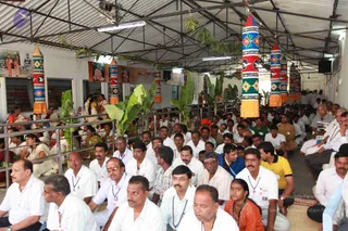 A large crowd of seated devotees fills a covered hall with a corrugated metal roof, many men in white shirts and white dhotis with lanyard badges, while women in saris sit at left. Tall decorative cylindrical banners in red, green, yellow and blue with floral and bird motifs hang from the ceiling, and potted banana plants stand among them. Framed printed pictures and posters line the back wall, and a loudspeaker is mounted high on the right. A steel railing separates the seating area from a rear aisle.