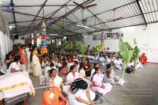 A photograph of a covered, open-sided hall with a corrugated metal roof and ceiling fans, where many people sit on a concrete floor in rows facing toward the left and the front of the room. Most seated attendees wear white or cream clothes, with some in orange, pink and maroon saris and salwar. A woman in an orange sari and a woman in a white and gold-bordered sari stand near the left, and a woman in a yellow-bordered outfit gestures toward the group. Tall painted cylindrical pillars wrapped in patterned fabric and green banana plants decorate the hall, and a rope-and-railing barrier separates the seated crowd from the front area. Framed photographs hang on the back wall, and a seated figure in orange robes appears at the far right.