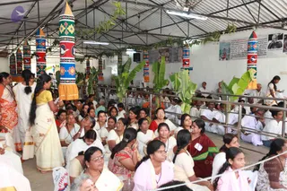 A photograph of a large open-sided pandal with a corrugated roof, decorated with tall painted cylindrical pillars in blue, green, red and yellow with floral and bird motifs, and strung with leafy garlands and potted banana plants. Rows of devotees sit on the floor behind metal railings, mostly women in white and cream sarees and salwar kameez, some in orange, red and pink. A woman in a cream saree with a long braid stands at left, and a man in white kurta sits at right. On the far right wall, framed photographs and printed panels hang beside the seated crowd. The scene is a seated congregation during a gathering.