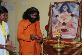 A photograph of a man in an orange saffron robe with a long dark beard and long wavy black hair lighting a small oil lamp held in his hands, beside a brass oil lamp on a draped red and gold cloth table. Behind him stands a young woman in a white coat with a yellow dupatta and bangles, watching. On the wall above the table hangs a framed portrait of a smiling man in orange robes seated in meditation, garlanded with white jasmine and red roses and marigolds. A small note on the wall reads 'Mind your mind before it minds you'. A signboard reads 'NO. 4 DENTAL'.