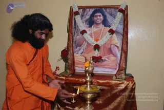 A photograph of a bearded man in an orange saffron robe lighting a small oil lamp with a flame on a brass lamp stand. Behind him on a table draped with gold-brown silk is a framed portrait garlanded with white jasmine and red roses, showing a smiling figure in orange robes seated in meditation pose before a tree trunk, with a rudraksha-style garland. A larger brass lamp with a yellow marigold sits in front. A small pink flower arrangement lies at the table's left side. The wall behind is pale yellow.