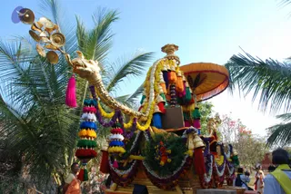 A decorated festival chariot, ratha, in a palm-lined outdoor setting under a clear blue sky. The vehicle has a golden serpent-headed prow with a fan of brass bells and cymbals at its front, and a golden canopy shaped like an umbrella with orange fringe. The chariot is draped with garlands of marigolds, purple and white flowers, tassels in red, blue, green and yellow, and hanging brass bells. Two or three onlookers stand near the chariot, one woman in an orange sari at the left edge and a few people in white and yellow at the right. The scene is a procession with a richly ornamented vehicle.