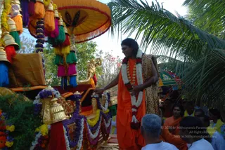A photograph of an outdoor festival procession beside a richly decorated palanquin or ratha draped with marigold and purple flower garlands, golden bells, colourful tassels and an orange canopy. A man with long swept-back hair, wearing an orange robe, a white and red flower garland, and a leopard-print shawl, stands at the side with his hand resting on the palanquin, while a few people in white and orange clothing sit or stand nearby. Palm fronds and a green tree frame the scene. Probable Swamiji is shown in the procession, identified by long swept-back hair, orange ceremonial robe, leopard-print shawl and flower garlands.