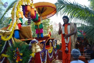 Outdoor procession scene beside a decorated ratha-style palanquin with a golden elephant head ornament, a large yellow fringed canopy umbrella, and tassels in red, green, blue and pink hanging from brass bells. Garlands of marigold and purple flowers wrap the vehicle. A man in saffron-orange robes, long swept-back hair and a leopard-print shawl, wearing white flower garlands and a gold necklace, stands at right waving his hand and smiling toward the crowd. Several people in white and orange clothing stand at the lower right, and palm fronds frame the top of the image.