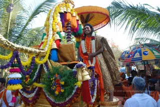 A decorated ceremonial chariot (ratha) draped with marigold and white flower garlands, purple and yellow tassels, and hanging bells, with a large orange-and-gold umbrella overhead. A man with long dark hair and a red tilak stands on the chariot in an orange-saffron dhoti with a leopard-print shawl over his shoulder, wearing white and red flower garlands and a rudraksha-style mala. Coloured parasols and devotees in white and orange stand along the procession route beside palm trees. a person: long swept-back hair, saffron clothing and a shawl, standing as the honoured figure on the procession chariot.