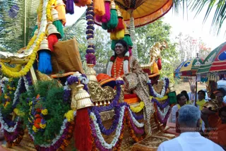 A decorated golden ratha procession vehicle, draped in garlands of purple, white and yellow flowers and hung with brass bells and multicoloured tassels, carries a smiling man seated on a golden throne. He wears an orange robe, a rudraksha mala and a white shawl printed with a leopard pattern. Overhead a yellow canopy and a multicoloured parasol shelter him. Devotees in white and orange stand beside the chariot, with a bald man in white in the foreground and more people behind the vehicle. Swamiji (probable): the seated man has long swept-back hair, orange clothing, rudraksha beads and a ceremonial golden necklace, so he is a probable depiction of Swamiji riding in procession.