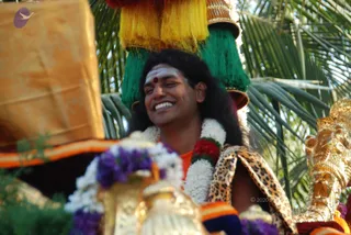 A smiling man with long dark hair, a white sacred ash line and red bindi on his forehead, sits in a procession. He wears white and jasmine garlands, a red flower garland, an orange garment and a leopard-print wrap. Above him rise a gold-trimmed headdress with green and yellow tassels. Behind him palm fronds fill the background, and a gilded deity figure with a crown is visible at right. A procession ratha is implied by the setting. Swamiji (probable) appears in this scene as the central figure, shown by long hair, orange clothing and rudraksha-style ceremonial adornment.