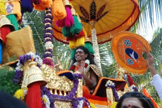 A photograph of a festival procession on a decorated golden chariot draped with garlands of purple and white flowers and hanging tassels in orange, green, pink and yellow. A smiling man with long dark hair, a tilak on his forehead, a red and white flower garland, and a leopard-print shawl over orange robes sits on a cushioned seat under a large yellow umbrella with a lotus motif, waving his right hand. Palm trees stand behind. A person at right holds up an orange fan-shaped ceremonial object with a bird emblem, and a partially visible woman with a red bindi appears at the bottom edge. a person: long swept hair, leopard-print shawl over orange robes and garlands fit the page's identification, so he is marked as in person.