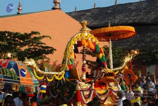 A photograph of a gilded ceremonial chariot procession outdoors beside an orange-washed building with a thatched roof. A golden ratha with curved dragon-headed finials is draped in yellow, orange, purple and white marigold garlands and hung with tasselled bells. An orange fringed umbrella shades a seated figure in orange and gold robes at the rear of the chariot, with a round orange fan or shield at the side. Around the chariot, crowds in white and yellow shirts and saris press close, and a striped multicoloured umbrella is held at the left edge.