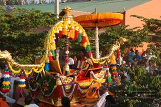 Outdoor procession scene of a richly decorated golden palanquin-style chariot with a carved gilded canopy shaped like serpent or makara heads, hung with rainbow tassels, bells, and garlands of marigolds and purple flowers. Beneath a large orange fringed parasol sits a figure with long dark hair wearing a saffron-orange garment, a white flower garland and a gold-embroidered shawl, seated on an orange cushioned throne. Around the chariot, white-clad attendants walk and devotees in white and colourful clothes line the route, some watching from behind green trees and a tiled-roof building. Several people are visible, including a man in white at the lower right and onlookers at the right edge.