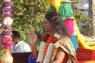 A man with long, swept-back dark hair rides on a decorated procession vehicle, waving his right hand and smiling. He wears a leopard-print shawl over an orange garment, with a white jasmine garland and a red-and-green floral garland around his neck, and a rudraksha-style ornamental necklace. Behind him rise tall tassels in yellow, green, pink and blue, and gold-trimmed decorative poles on the chariot. On the left, a purple and pink floral garland hangs from a gold pillar, and a man in white stands in the background among green leaves. Swamiji (probable) is the central figure, identified by long hair, orange clothing, ornate garlands and a procession setting, with the ceremonial vehicle indicating a ratha procession.