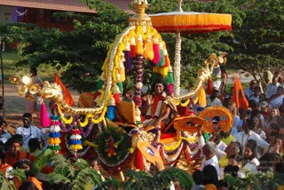 A golden ornate chariot procession moves along a path lined with crowds. The gilded ratha is decorated with yellow and marigold garlands, purple and white floral strands, and dangling bells and tassels in red, orange, blue and green, with a yellow and red fringed canopy overhead. A man with long dark hair, wearing a crimson and gold ceremonial garment with a white garland, sits on the raised seat and raises one hand in greeting. Around the chariot, devotees in white and saffron-orange clothing walk and watch, some holding orange flags. Green trees and a red-roofed building sit in the background. Printed copyright text is not included.
