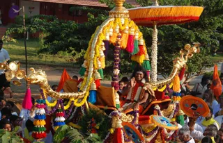 A golden ceremonial chariot with a dragon-headed prow decorated with garlands of marigolds, white flowers, and strings of gold bells is carried through a procession. A man seated in the chariot wears an orange-red garment with a gold-embroidered shawl, a white floral garland, and a red and gold mantle, and he raises his hand in greeting with a broad smile. An ornate orange umbrella with red fringe shelters the chariot. Colourful tassels in pink, green, blue, and yellow hang from the structure. Orange and blue decorated fans are held by attendants, and a crowd of devotees in white clothes stands along the roadside with a tree and red-roofed building behind. a person appearance cues are present: the seated figure has long swept-back hair and ornate attire, so this is an appearance-based candidate in person.