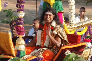 A smiling man with long dark swept-back hair sits on an ornate golden palanquin decorated with purple and white flower garlands and green, pink and yellow tassels. He wears an orange robe, a red and white flower garland, a tiger-print shawl draped over one shoulder and a white tilak with a red dot on his forehead, and raises one hand in greeting. Behind him stand several people in white clothes, including a shaved-headed man in white with an orange cord, and a woman in the background. Dry ground and trees form the setting. Swamiji (probable): the seated figure shows long swept-back hair, saffron robes and ceremonial gold and floral adornment, as a procession participant on the palanquin.
