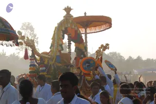 A hazy outdoor procession at golden hour shows a richly decorated ceremonial vehicle carried through a crowd. The vehicle is draped with yellow, orange, red and white garlands and fabric, topped by an orange fringed canopy and a tall golden kalasha-like finial, with colourful tasselled tassels hanging at its sides. Seated on the vehicle is a figure in orange and red cloth whose face is not clearly visible; a murti or devotee is not distinguishable. Flanking the vehicle, attendants hold round decorated fans and ceremonial staffs. The foreground crowd is dominated by men and women in white dhotis, shirts and saris, many with raised hands, with palm trees and tents in the background.