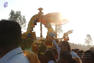 Photograph of an evening procession in warm backlight. A decorated palanquin or ratha with a canopy of orange and yellow tassels, marigold garlands, and a golden umbrella-shaped top is carried forward. A woman in white, with hair tied back, raises a ceremonial fan or chamara-like object toward the canopy. Around her, devotees in white and orange clothing crowd the road, many seen from behind, some partially visible in the foreground. Trees and bright sunlight fill the background. No clearly identifiable a person figure is visible on the palanquin.
