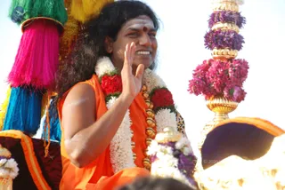 A smiling man in an orange sleeveless sadhu garment and a red bindi waves from a decorated procession vehicle. He wears long layered garlands of white jasmine and red flowers with gold beads. Behind him stand tall pillars of purple and pink flowers on gold bases, and multicoloured tassels of pink, blue and green hang at left. The vehicle carries orange and maroon cloth edged with gold trim. Bright sunlight fills the background.