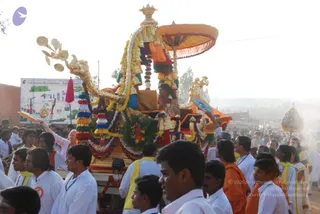 A decorated golden procession vehicle, a ratha-style palanquin, moves through a crowd during an evening procession. The structure is draped with garlands of marigolds and green leaves, colourful tassels in red, yellow, green and white, and an orange canopy with fringe. A figure seated on the vehicle wears an ornate dark garment with gold trim and a flower garland. Ceremonial gold fans and a curved gold standard stand at the front. Around it, many men in white shirts and white dhotis, some wearing yellow or orange shawls and lanyards, walk closely beside the vehicle. A roadside billboard reads Nithyananda Dhyanapeetam - Bidadi Ashram. Hazy dusty light fills the background.