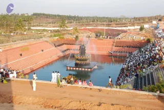Aerial photograph of an outdoor temple tank with a large black Shiva linga on a square platform in the centre, decorated with a coloured garland band and water jets rising around it. Tiered red-orange stone steps line the sides of the tank, and dense crowds of devotees in white and colourful clothes fill the stands on the right. Several people in white dhotis and shirts stand along the edge of the tank and on the dirt path, with a few women in pink and green saris nearby and a flower-decorated offering tray at the bottom left. Palm trees and pine trees surround the grounds, and parked cars line the right edge.