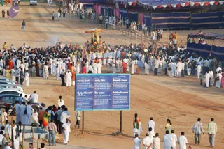 Wide outdoor photograph of a crowded procession on a dusty open ground lined with blue-and-white tent canopies and bamboo barriers. At the centre, a decorated palanquin or ratha carrying a gilded canopy and a small ornate figure moves forward, flanked by umbrellas and attendants in white. Dense crowds of devotees in white dhotis, kurtas and sarees fill the road, with some in orange and saffron clothing. In the foreground a large blue signboard with multilingual text describes the Ananda Linga, Vaidya Sarovar and a healing site. Parked cars line the left edge, and a faint smoke plume rises near the procession.