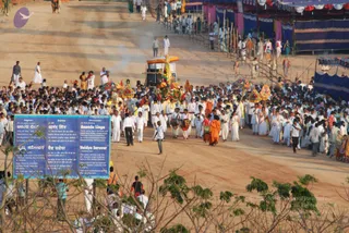 A large outdoor procession moves down a dirt road lined with blue and white tents and bamboo barricades. At the centre, a decorated palanquin or float carries a tall garlanded structure topped with a golden canopy, with a smoke trail rising behind it. Walking around it are men and women in white ritual clothes, several carrying drums, and a few in orange or saffron robes. Large parasols in red, yellow and green lead the group. A blue signboard in the foreground reads Ananda Linga and Vaidya Sarovar with Healing Lake text in several scripts. Dense crowds of white-clad devotees fill the roadway and sides, with onlookers standing among the tents.
