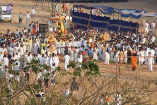 A large outdoor procession on a dusty ground, seen from an elevated angle. Crowds of people in predominantly white clothes walk around a decorated palanquin or ratha with a gold and red canopy and ornate golden top, carried forward by a group of men in white dhotis and white shirts. Several people in saffron-orange garments walk among them, and a drummer beats a drum at the front. Blue and white tented shelters with scalloped edges line the path on the right, and a white van drives at the upper left. Bare branches frame the lower foreground. The crowd is dense and extends beyond the frame.