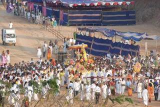 Aerial view of a large outdoor procession on a dirt road lined with blue-and-white canopy tents and bamboo fencing. A decorated palanquin with a golden canopy, garlands and umbrellas is carried through a dense crowd of devotees, most dressed in white with some in yellow and orange. Drummers in orange and white stand near the front, and a white van drives along the left edge. Umbrellas with fringed, colourful canopies flank the palanquin. Hills rise in the background.