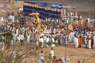 A large outdoor procession of many people dressed in white moves across sandy ground past blue tarpaulin-roofed stalls and bamboo poles. At the centre, a decorated palanquin or float with a yellow canopy, garlands and gold ornaments is carried forward. Men in white shirts and dhotis, and women in white saris, walk alongside; several figures in orange or saffron robes walk near the float. Drummers carry drums, and colourful decorated umbrellas and banners flank the procession. Trees and bushes frame the lower foreground. No one can be clearly identified as a person from appearance; the figures visible are a crowd of devotees and attendants.