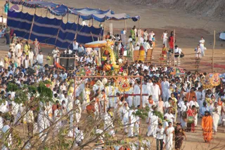 Outdoor evening procession on sandy ground with a large blue-and-white striped tent canopy behind. A decorated palanquin with a yellow canopy and garlands carries a deity murti at its centre, pulled along by a red rope, surrounded by a dense crowd of devotees mostly dressed in white, with some in saffron, green and pink saris. Several people carry colourful umbrellas and drummers play drums at the lower right. At the far lower right, a figure in an orange-saffron robe walks among the crowd, possibly a sannyasi or a person-like figure, but appearance is not distinctive enough to confirm identity. Background shows scattered onlookers and tables.